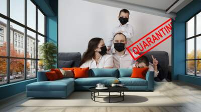 young couple and two children are sitting in a room on a gray sofa in black virus masks, red inscription quarantine, concept of quarantine measures during the period of the disease epidemic Wall mural