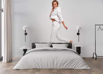 Beautiful blonde woman jumping in the air in white tracksuit isolated on gray background Wall mural