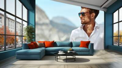 Portrait of handsome man wearing elegant white shirt and sunglasses, standing near the lake in the alps Wall mural