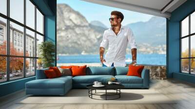 Portrait of handsome man wearing elegant white shirt and sunglasses, standing near the lake in the alps Wall mural