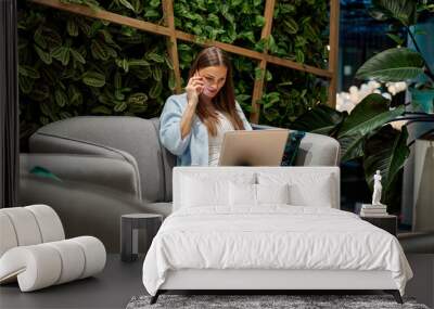 Young female woman using laptop and phone, communicates on internet with customer in home garden/greenhouse.Cozy office workplace. Wall mural