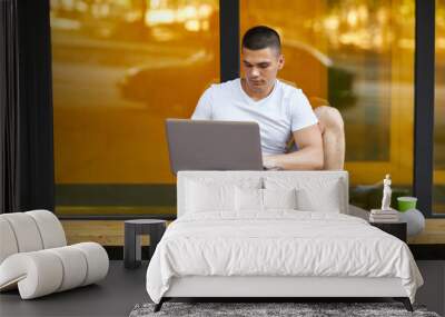 Young man sitting on the park bench with laptop on his lap Wall mural