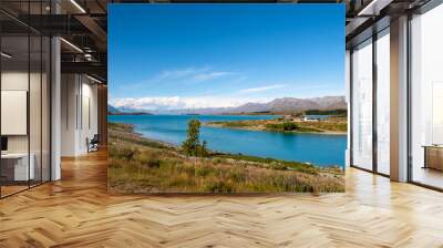 A scenic lakeside view with a charming stone church, surrounded by distant mountains and a nearby parking area. The bright blue sky and clear waters enhance the tranquil atmosphere, making it a pictu Wall mural