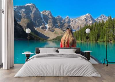 Moraine lake canoeing. The blonde woman in red life jacket sitting in the canoe with the beautiful view on the Valley of the Ten peaks and the turquoise lake surrounded by pine trees. Blue sky. Wall mural