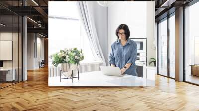 a young woman in glasses and in a blue shirt  works at home at a laptop as a coach in front of the kitchen in a bright interior, a concept of a workplace and remote work at home Wall mural