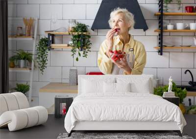 beautiful elderly gray haired senior woman cook in cozy kitchen with fresh organic vegetables, tomatoes, cabbage, lettuce, cucumbers on table cooking healthy vegetable salad, healthy food active life Wall mural