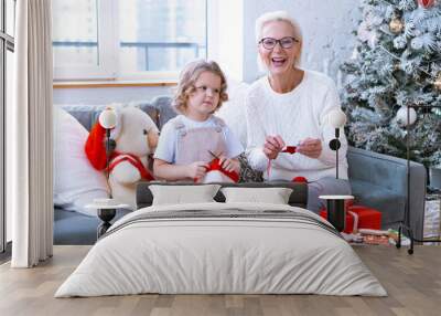 Christmas family grandmother and granddaughter sit on sofa near Christmas tree and preparing handmade New Year gifts, pretty girl and active happy senior woman knitting and sewing toys in apartments Wall mural