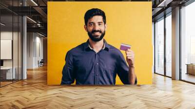 Portrait of a smiling bearded man showing credit card isolated over yellow background Wall mural