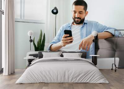 Smiling man sitting on couch at home using laptop and cell phone Wall mural