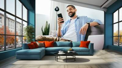 Smiling man sitting on couch at home using laptop and cell phone Wall mural