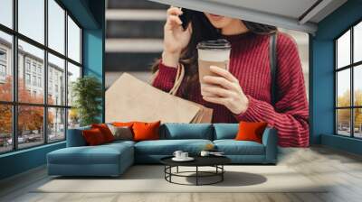 Young pretty woman blogger with smartphone, cup of coffee and shopping bags in city mall Wall mural