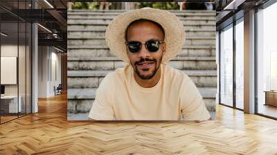 Young attractive dark-skinned male in trendy straw hat and sunglasses sitting on the stairs. African man hipster treveler relaxing at city. Leisure and rest at summer day Wall mural