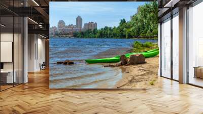 Green kayaks on the lake. A beach with yellow sand and two green kayaks. Coastline against a cityscape Wall mural