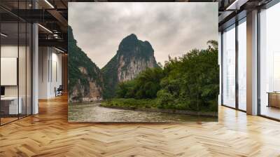 Guilin, China - May 10, 2010: Along Li River. Landscape of 2 karst mountains with vertical brown-black cliffs under rainy cloudscape right around bend in river with green trees on shore. Wall mural