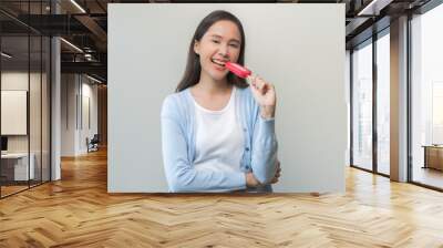 Cheerful, happy positive asian woman wearing casual with brunette hair, enjoy eating delicious popsicle, girl holding cold dessert, frozen stick with strawberry ice cream tasty isolated on background. Wall mural