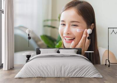 Facial beauty skin health care, smile of cute asian young woman looking at mirror, hand applying moisturizer lotion on her face, female putting cream treatment before makeup cosmetic routine at home. Wall mural