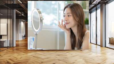 Fresh healthy skin, beautiful smile of asian young woman, girl looking at mirror, applying moisturizer on her face, putting cream treatment before make up cosmetic routine at home. Facial Beauty. Wall mural