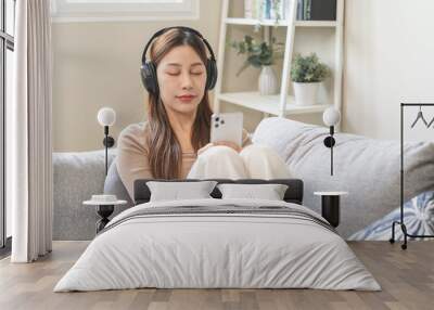 Relaxing asian young woman using smart phone listening to music, enjoy song or watching videos, podcast on mobile with white wireless headphones sitting on sofa, couch at home, chill out and leisure. Wall mural