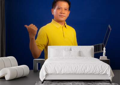 Portrait of smiling middle-aged asian man in casual yellow polo shirt holding laptop isolated on blue background in studio, looking happiness and attractive professional leadership Wall mural