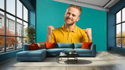 Man in yellow wear is very glad and happy, he shows yes gesture of victory, guy achieved result, goals. Surprised excited happy guy on blue background Wall mural