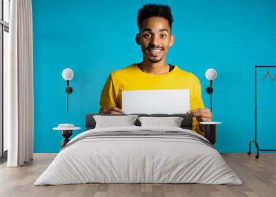 Portrait of young handsome african american man holding white empty paper blank on blue studio background. Copy space. Positive guy with sign Wall mural