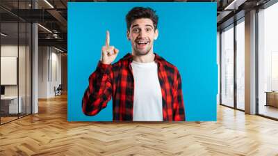 Portrait of young thinking pondering man in red having idea moment pointing finger up on blue studio background. Smiling happy student guy showing eureka gesture. Wall mural