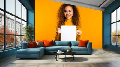 Pretty curly woman holding white vertical a4 paper poster. Copy space. Smiling trendy girl on yellow background. Wall mural