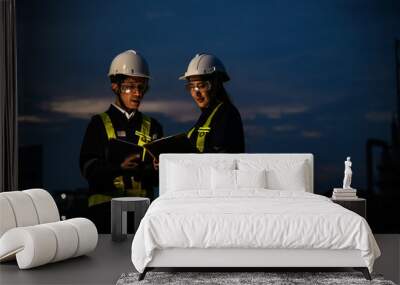 Two engineers, wearing helmets and reflective vests, work on a laptop at night at an industrial site. Industrial plant and structures are illuminated in the background. Wall mural