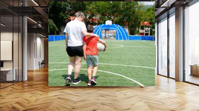 Mother and son walking arm in arm on a sunny soccer field with a ball under the mother arm. Wall mural