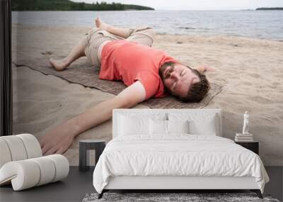 Bearded man practices yoga, he performs an exercise lying on a carpet, on a sandy beach by the lake, on a summer day. Wall mural