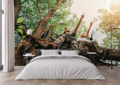 Older sister and younger brother sit together on tree in forest and point their hands against background of green trees and nature. Childhood and friendship. Two children climb on branch. Low angle Wall mural