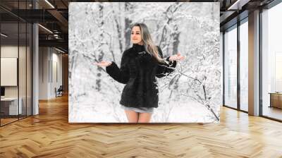 Beautiful young girl in fur coat and dress with bare legs in winter in a snowy forest catches snow with her hands Wall mural