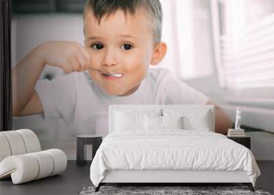 Cute boy in the kitchen eating yogurt from a white yogurt container, a place for advertising Wall mural