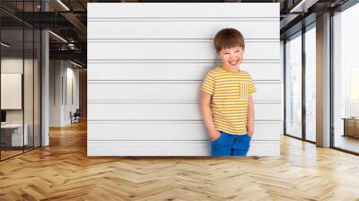 Portrait of smiling boy on white background with stripes. Cute kid stands hands in pockets near light grey wall. Child in yellow t-shirt and blue jeans laughs joyfully. Wall mural