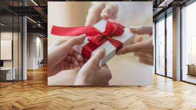 A small present is being passed by a couple from one to another. White little giftbox with a red ribbon in the hands. Valentine's day and winter holidays concept. Wall mural