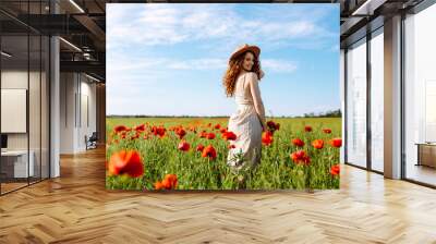 Beautiful girl posing in a poppy field. A woman with a hat stands in the middle of flowers. The young girl smiles. Happiness concept. Relaxing on summer poppy flowers meadow. Wall mural