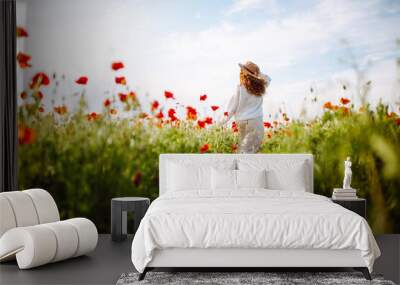 Girl in a hat with long curly hair posing in a field with red flowers. Summer landscape. Warm colors. Woman walking through a poppy field. Young girl in the spring flower garden. Wall mural