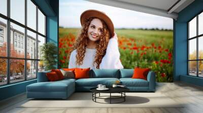 Portrait of a beautiful young woman in a poppy field. Happy curly girl in a field with flowers. The girl smiles in the middle of meadow with beautiful red flowers. Happiness concept. Spring landscape. Wall mural