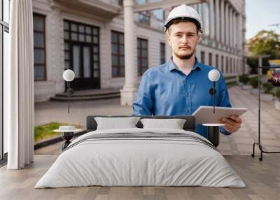 Young architect man wearing contractor helmet holding project paper plan and tablet against the background of the object. Portrait of civil engineer. Wall mural