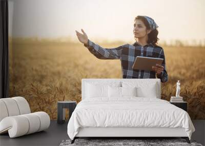 Young woman farmer stands in the wheat field looking for the progress of the new crop growth. Female farm worker with a tablet is checking the wheat quality before harvesting. Smart farming concept. Wall mural