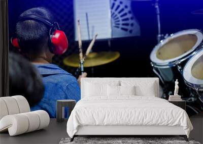 Asian boy put blue shirt and red headphone to learning and play drum set with wooden drumsticks in music room. Teacher teach boy to pay drum set. The concept of musical instrument, Selective focus. Wall mural