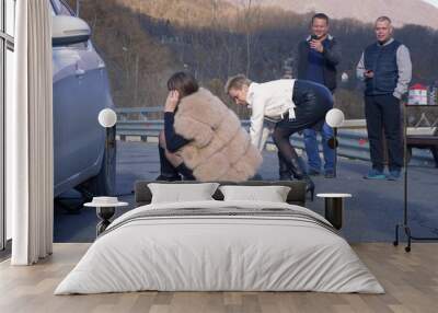 two girls changing a car wheel on the side, and the men watching from the side Wall mural