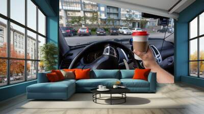 Closeup of a young man car driver drinking coffee, hand holding a paper white coffee Cup with a red lid in the background steering car dashboard blurred Parking background. Wall mural