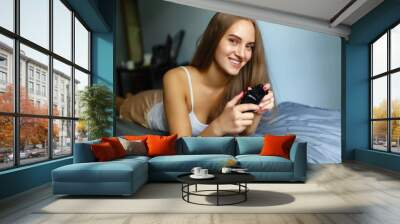 Girl gamer playing with a wireless controller, looking at the screen in front. Young girl eSports player smiling and enjoying the victory with gamepad in hand, playing video games on the console. Wall mural