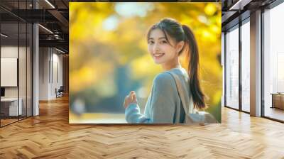 A cheerful young woman smiles while wearing a backpack in a sunny autumn park, surrounded by golden leaves and soft light. Wall mural