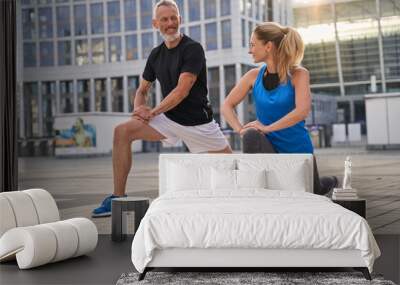 Active sporty middle aged couple, man and woman stretching legs, preparing for running together in the city on a summer day Wall mural