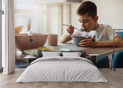 Caucasian small teen boy eating cereals with milk and drinking tea at table in morning. Wall mural