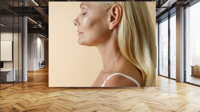 Cropped portrait of natural mature blonde woman in white underwear posing with eyes closed, standing isolated over beige background Wall mural