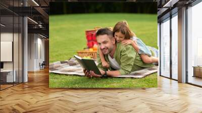 Curious little girl distracting, playing with her dad when he reads a book while lying on a blanket. Happy father and his daughter having a picnic in the park Wall mural