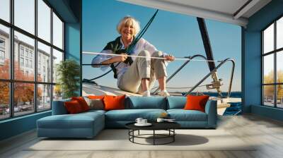 Enjoying sea trip. Happy senior woman sitting on the side of a sail boat or yacht deck floating in sea, looking at camera and smiling. Clear blue sky background Wall mural